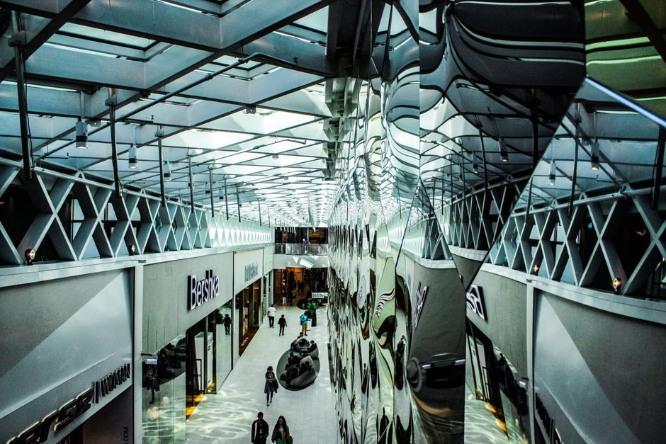 Inside a modern shopping mall with high glass ceilings and metal framework, the image shows a spacious corridor lined with retail stores, including Bershka and other fashion outlets. The polished, ref