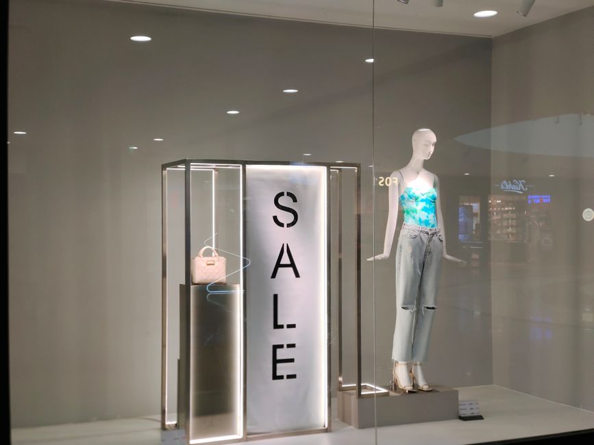 Inside a modern shopping mall with high glass ceilings and metal framework, the image shows a spacious corridor lined with retail stores, including Bershka and other fashion outlets. The polished, ref